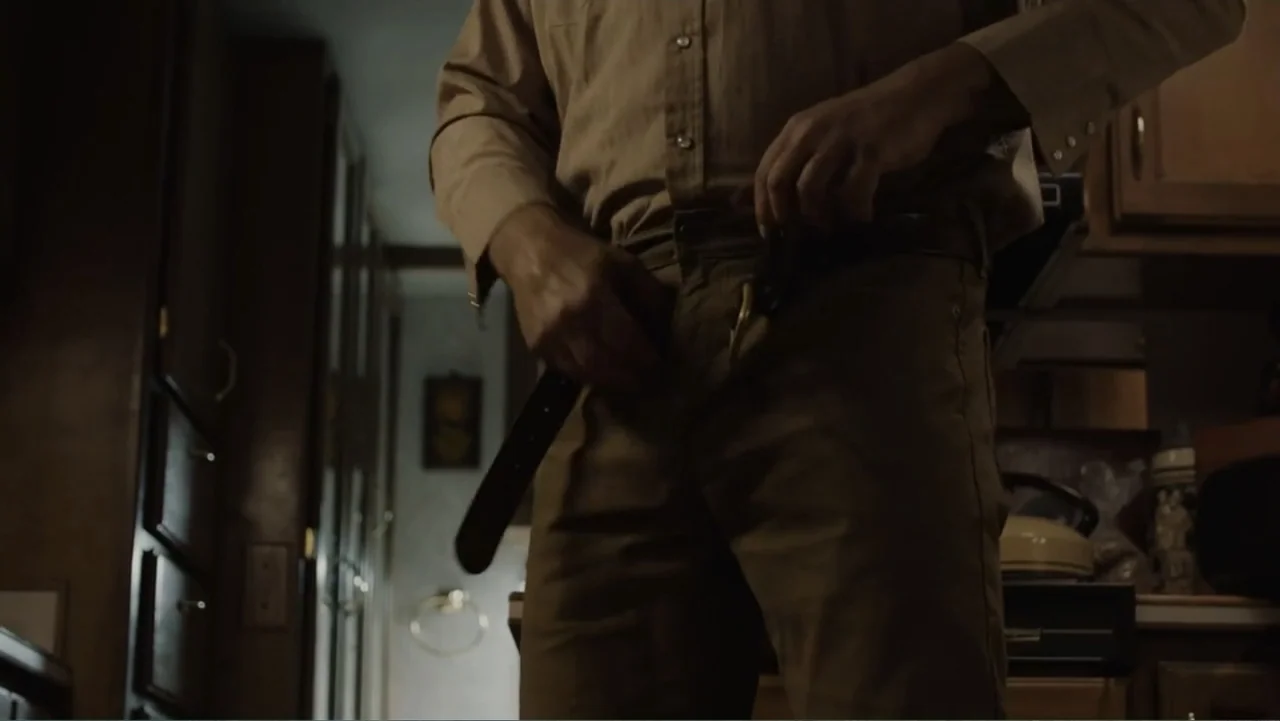 The Cleaner (2021) movie still: medium — A dimly lit interior of a compact home space featuring dark cabinets and appliances. A man wearing a tan…