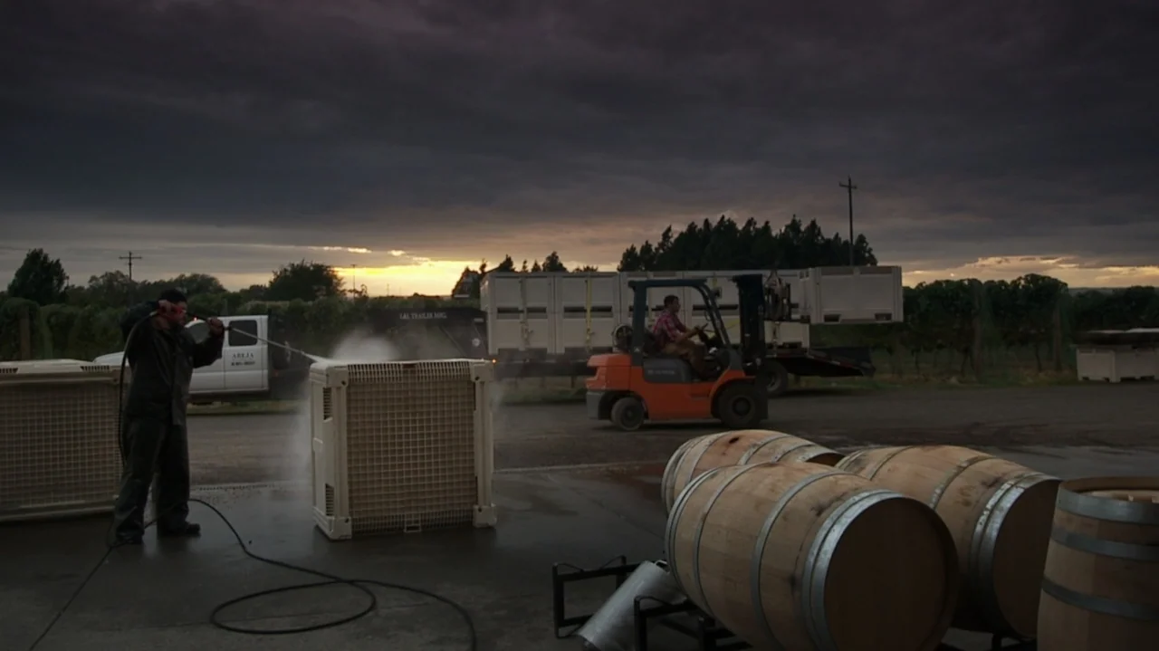 Somm: Into the Bottle (2015) movie still: wide — An outdoor winery area features wooden barrels in the foreground and equipment on asphalt. A worker…