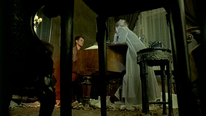 Almanac of Fall (1984) movie still: wide — A dark, cluttered room contains a wooden piano and an ornate pedestal side table. A man sits at the piano…