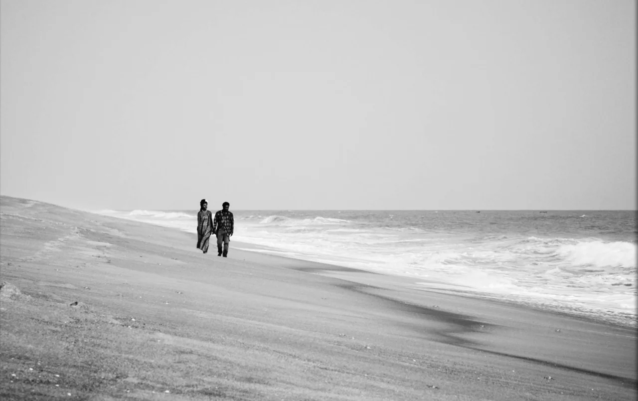 Mami Wata (2023) movie still: wide — A vast sandy coastline extends toward the horizon under a pale, overcast sky. A man and a woman walk…