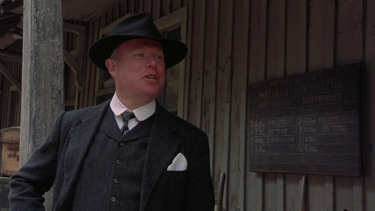 Matewan (1987) movie still: medium — A wooden train station wall features a painted black passenger train bulletin board. A man in a dark wool suit…