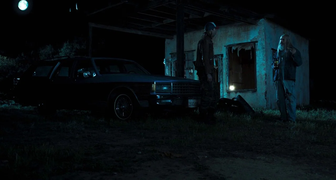 3 From Hell (2019) movie still: wide — A dilapidated, partially ruined single-story structure stands at night under a full moon. Two men stand…