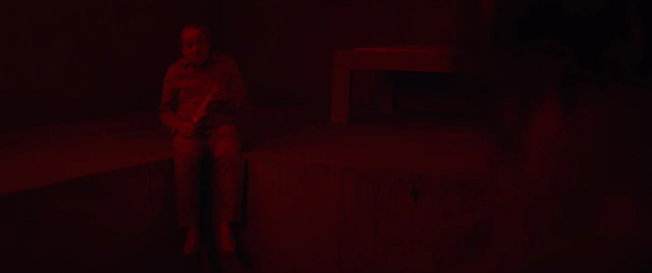 The Platform (2019) movie still: wide — A stark room with a raised concrete platform and a small table bench. An older man sits alone on the edge of…