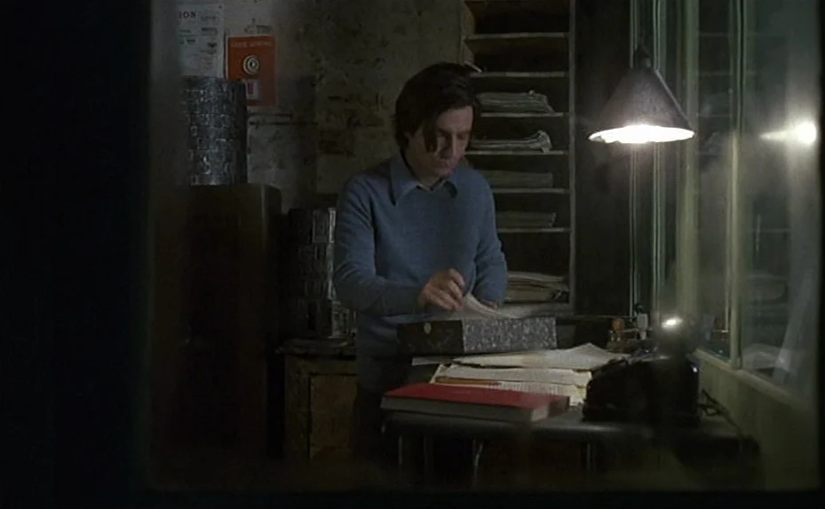 Love on the Run (1979) movie still: medium — A dimly lit room contains stacked shelves and a desk piled with loose papers. A man with…