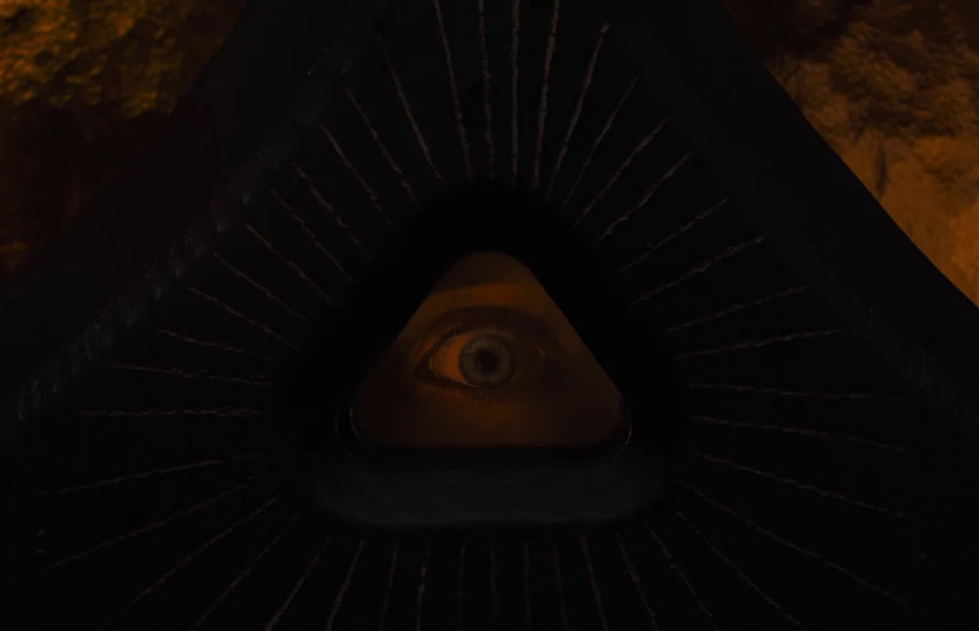 Gretel & Hansel (2020) movie still: closeup — A human eye peers through a triangular hole set into a dark, textured surface. The surrounding…