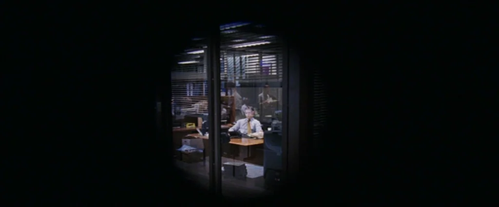 The Departed (2006) movie still: wide — A dark, empty workplace features a single illuminated office space behind glass panels. A man sits at his…