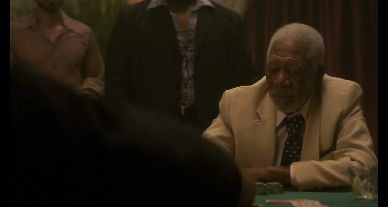 The Poison Rose (2019) movie still: medium — A dimly lit room features a green poker table where several men are gathered. An elderly man in a beige…