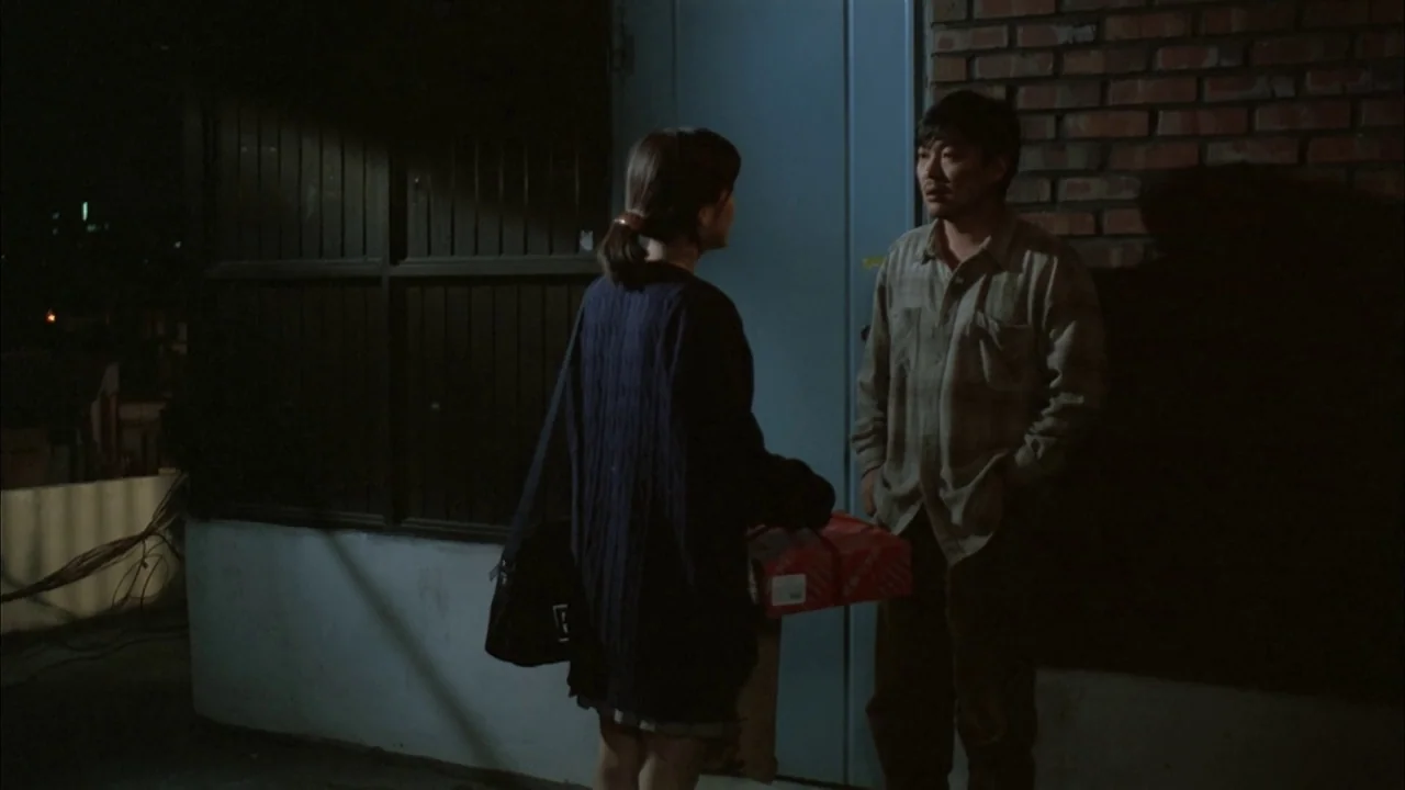 The Day a Pig Fell Into the Well (1996) movie still: fullbody — A night exterior showing a dark alleyway next to a red brick building. A woman…
