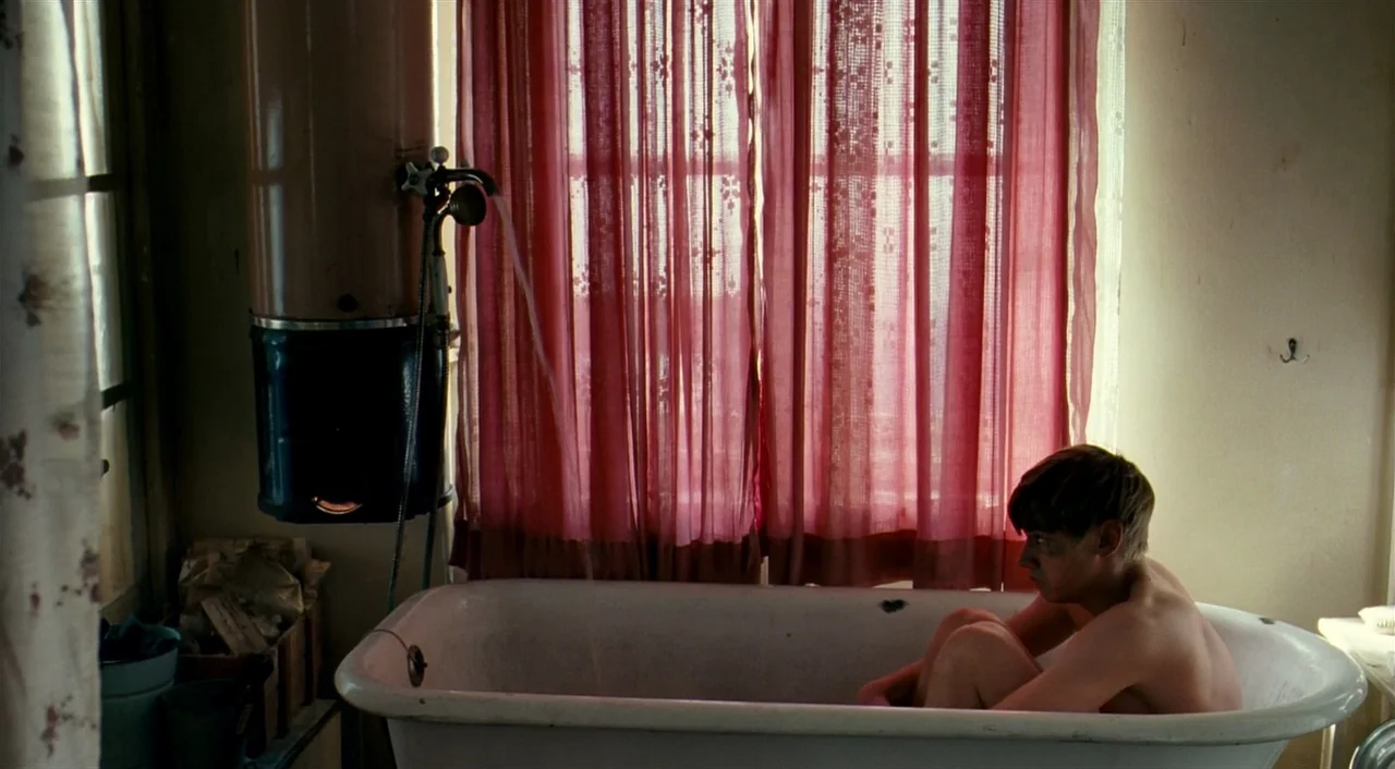 The Reader (2008) movie still: medium — A small bathroom features a vintage tub and a water heater near the window. A young boy sits inside the…