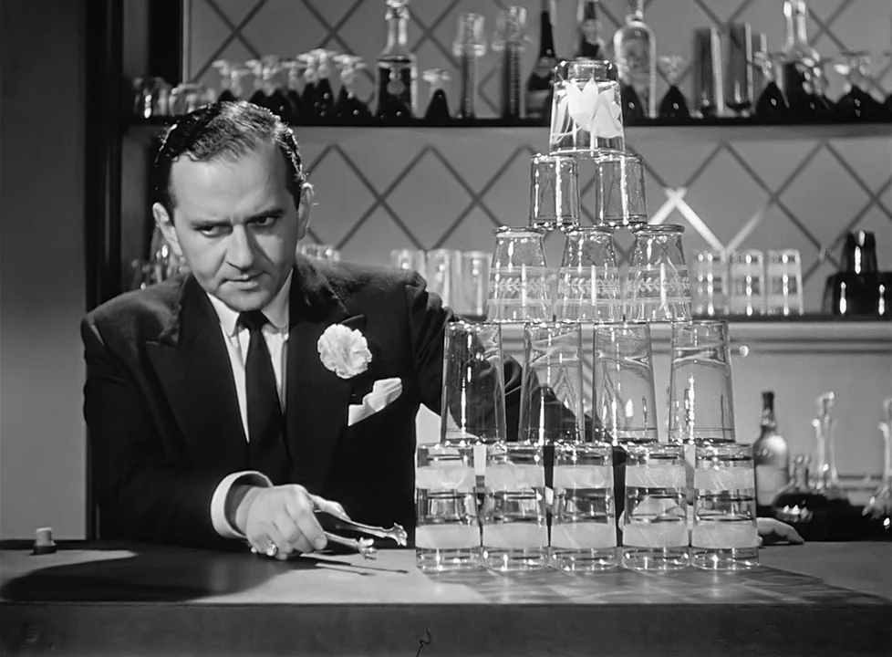 M (1951) (1951) movie still: medium — A man in a tuxedo stands behind a bar with a diamond-patterned mirror background. He holds silver ice tongs…