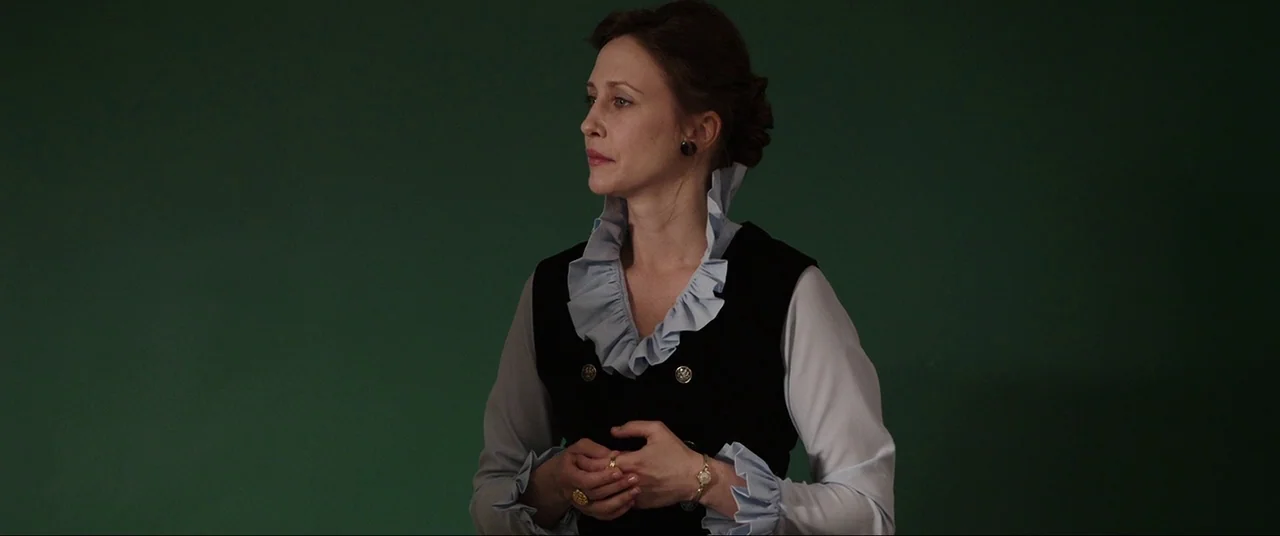 The Conjuring (2013) movie still: medium — A woman stands centered against a flat matte green studio backdrop. The woman keeps her hands clasped…
