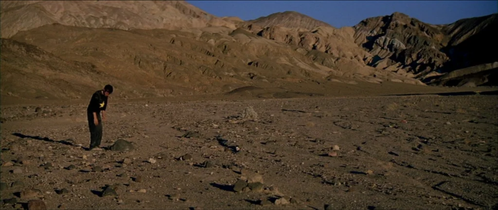 Gerry (2002) movie still: wide — A vast, arid desert landscape features dry soil, scattered rocks, and towering barren mountains. A young man stands…