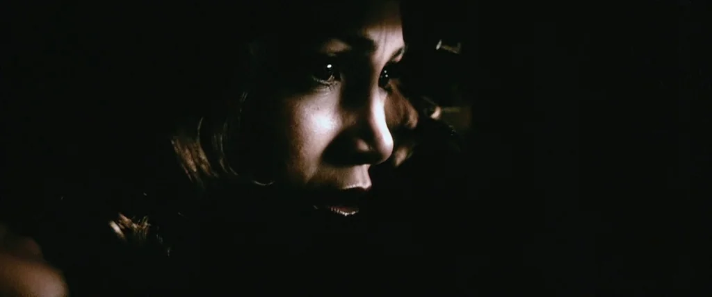 The Cell (2000) movie still: closeup — A person's face emerges from complete darkness into a narrow strip of light. The woman remains still with her…