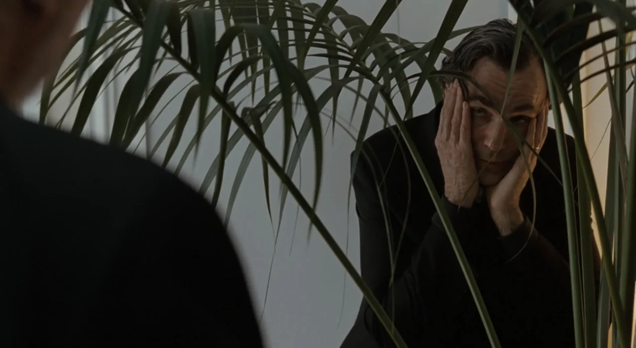 The Great Beauty (2013) movie still: medium — A bright office interior with a tall indoor plant partially obscuring the view. A man in a black suit…
