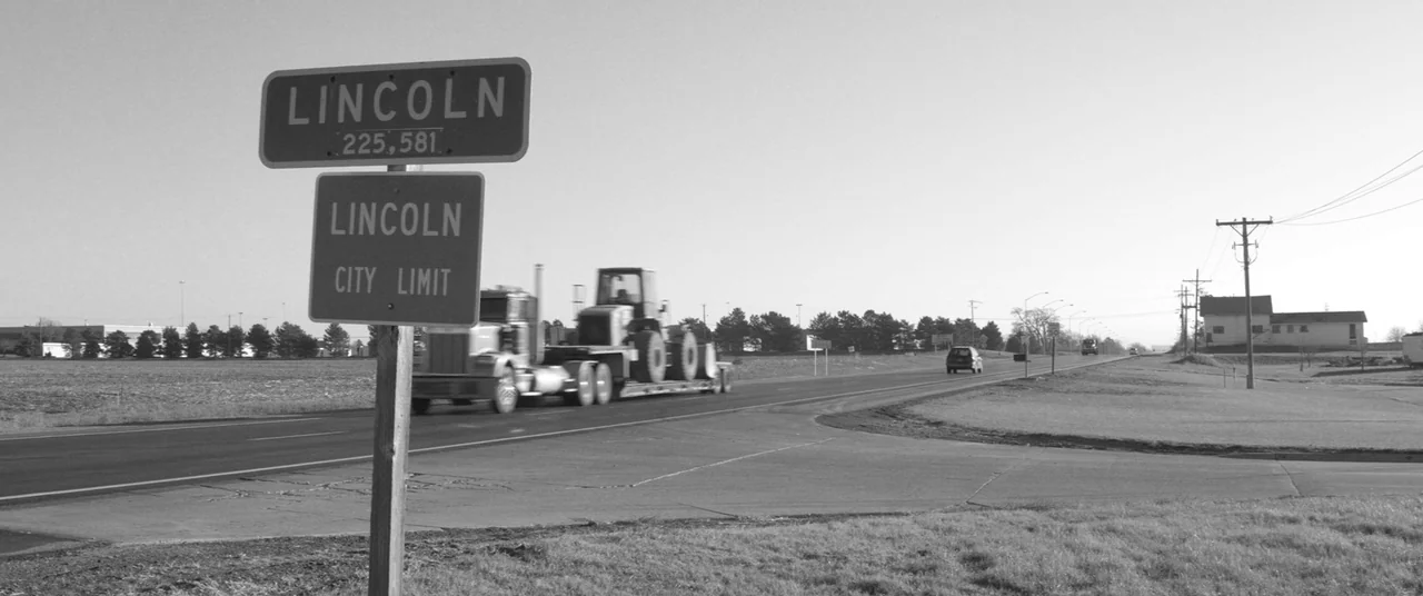 Nebraska (2013) movie still: wide — A highway stretches through a flat, rural landscape with a city limit sign. A semi-truck carrying construction…