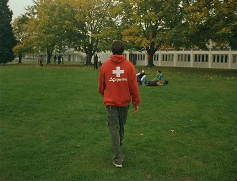 Elephant (2003) movie still: fullbody — A green grassy lawn sits in front of a white multi-story building with trees. A person in a red hoodie walks…