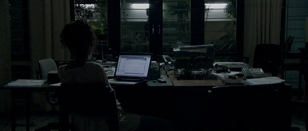 A Mighty Heart (2007) movie still: medium — A darkened room features a desk cluttered with papers and electronic equipment at night. A woman sits in…