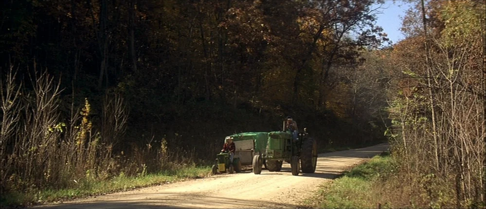 The Straight Story (1999) movie still: wide — A rural dirt road passes through dense deciduous woods with autumn-colored leaves. Two men operate…