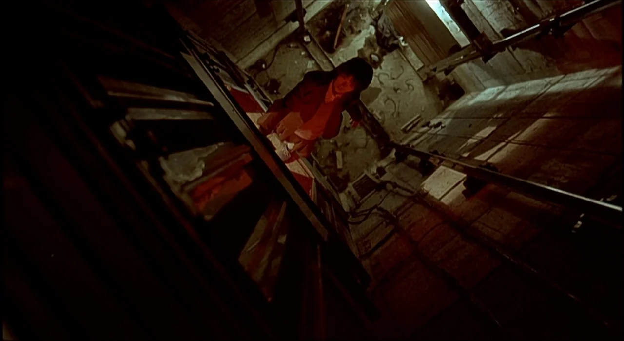 Voice (2005) movie still: wide — An industrial elevator cage with steel frame tracks extends into a dark, debris-filled shaft. A man in a dark…