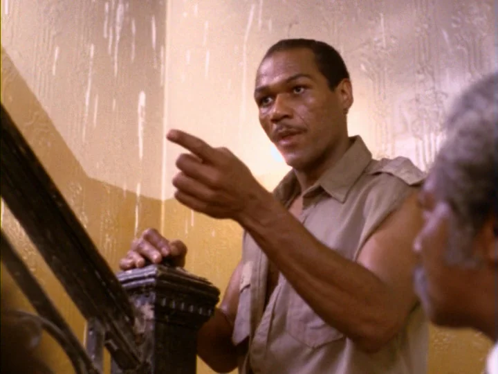 Tenement (1985) movie still: medium — A stairwell with beige walls featuring patchy paint and a dark wooden banister. A man in a tan shirt gestures…