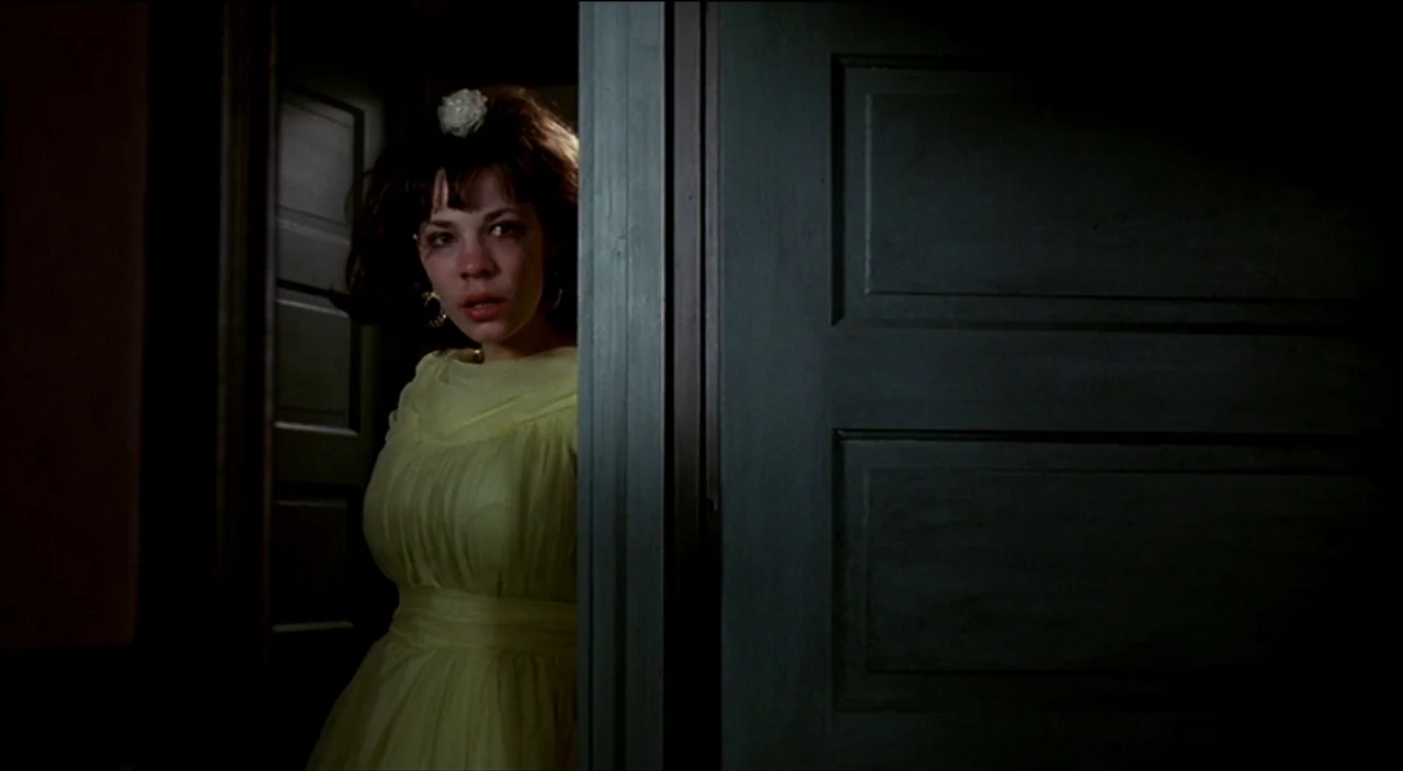 Dogfight (1991) movie still: medium — A woman stands in a partially open doorway within a dimly lit home. She wears a yellow dress and peers through…