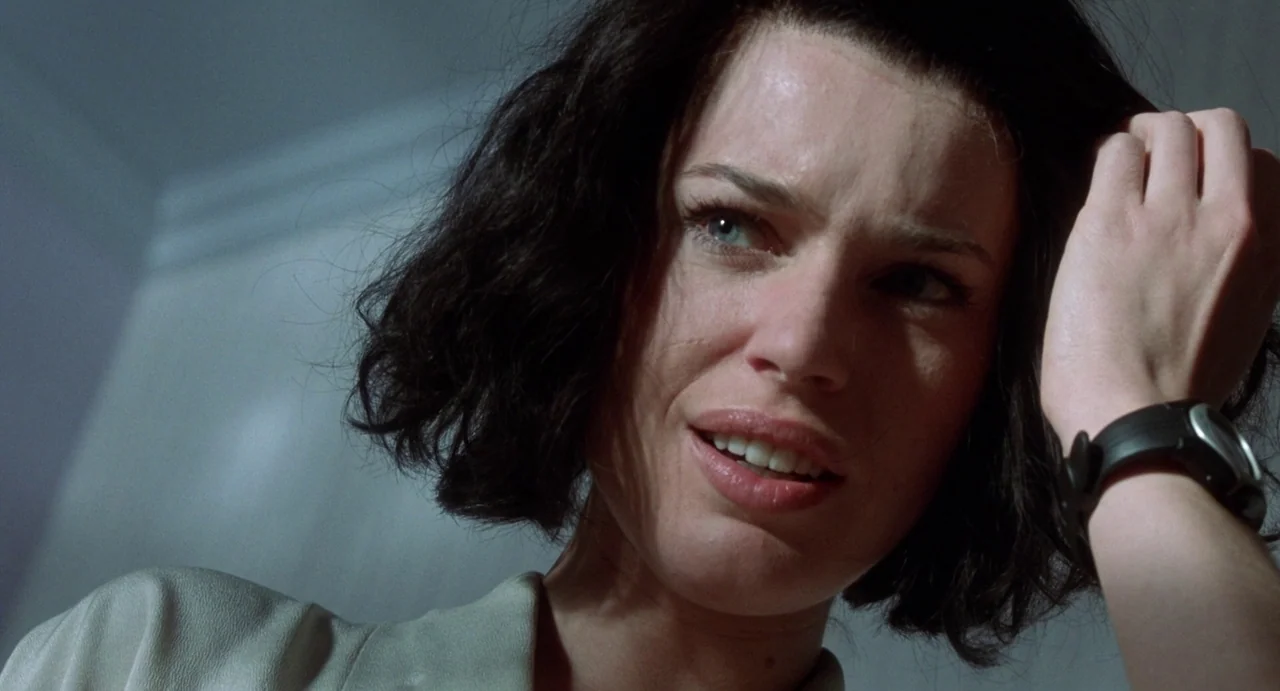 Femme Fatale (2002) movie still: closeup — A close-up shot focuses on a woman indoors with a plain light-colored wall behind. She is looking down…