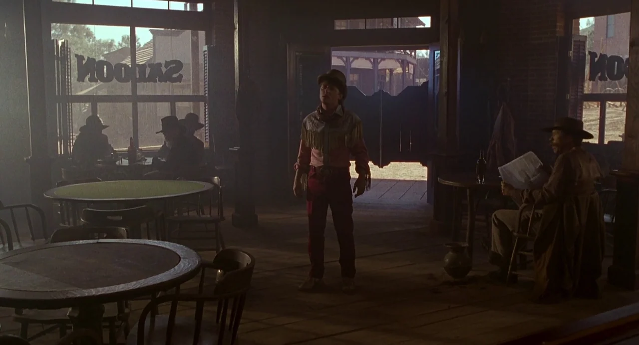 Back To The Future Part III (1990) movie still: fullbody — A dusty saloon interior with wooden tables and chairs under dim natural lighting. A man…