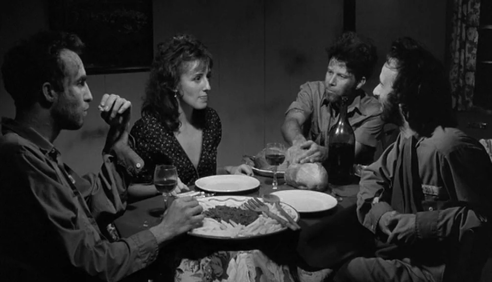Down By Law (1986) movie still: medium — A dimly lit room features four people gathered around a small circular dining table. The group eats from a…