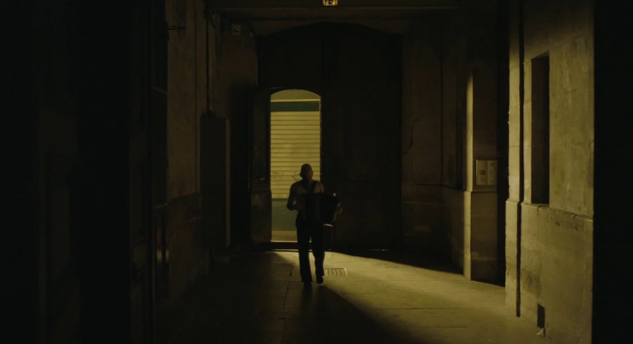 Holy Motors (2012) movie still: wide — A dark stone passageway with a bright rectangular opening at the far end. A man walks through the portal…