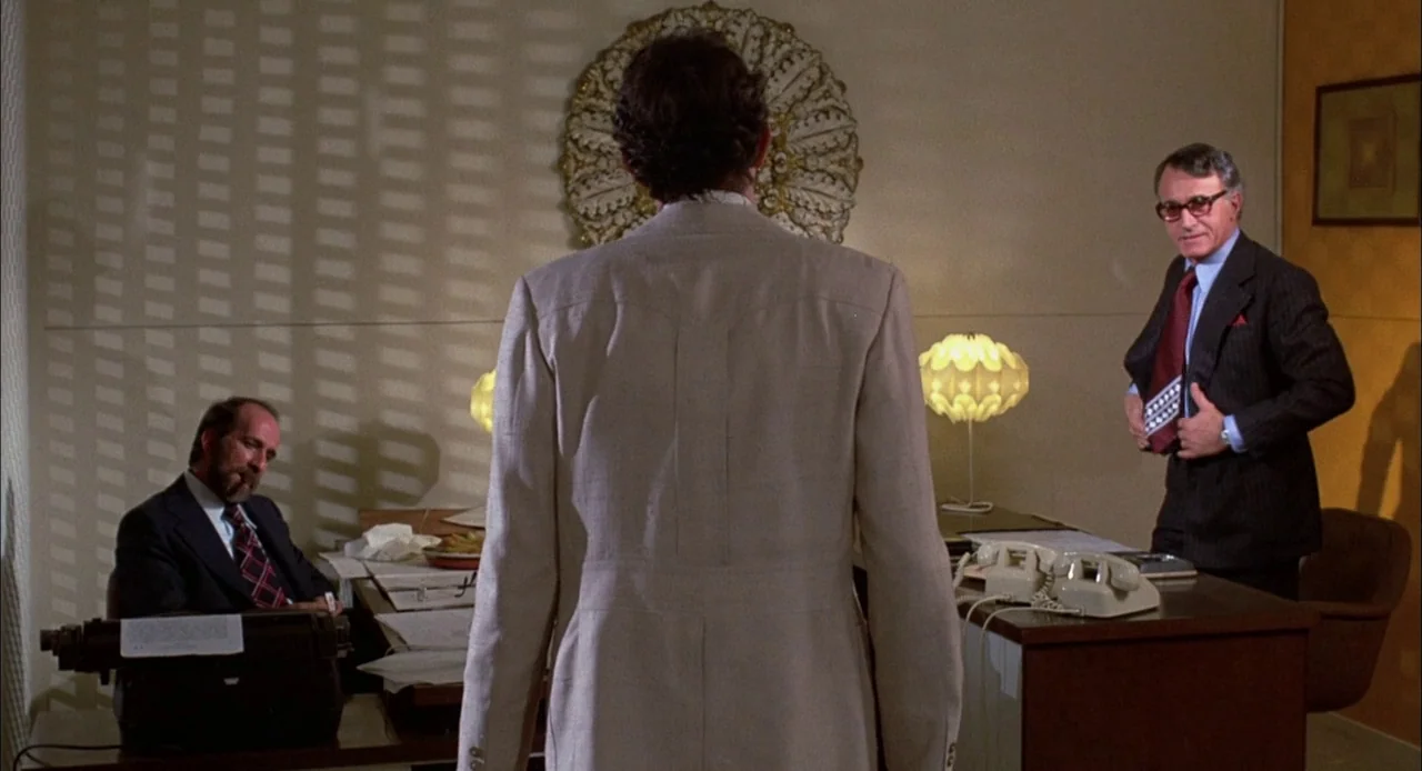 Bring Me The Head of Alfredo Garcia (1974) movie still: medium — A 1970s office interior with desks, a typewriter, and wood-paneled or papered…