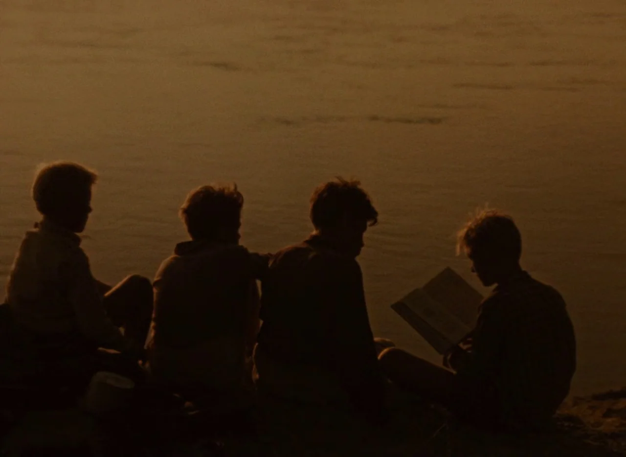 A Journey to the Beginning of Time (1955) movie still: medium — Four boys sit closely together on a dark riverbank at sunset. One boy holds an open…