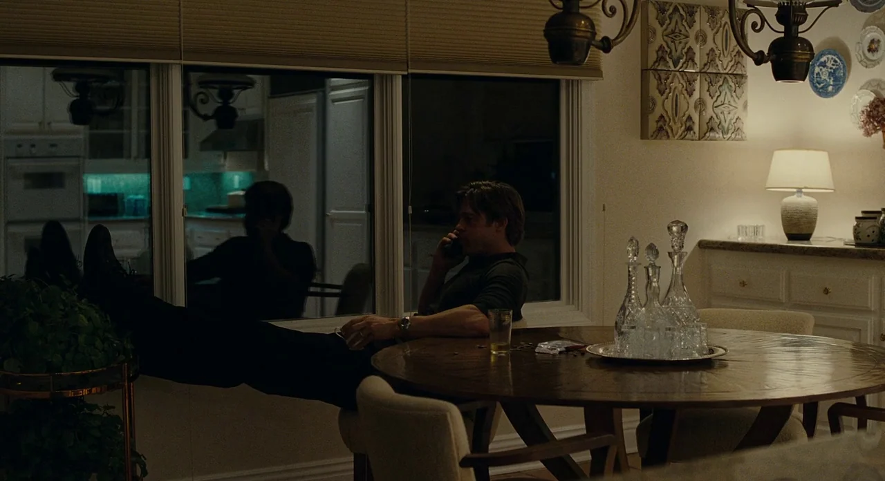 Moneyball (2011) movie still: medium — A quiet dining area features a wooden table with crystal decanters and a lamp. A man sits at the table,…