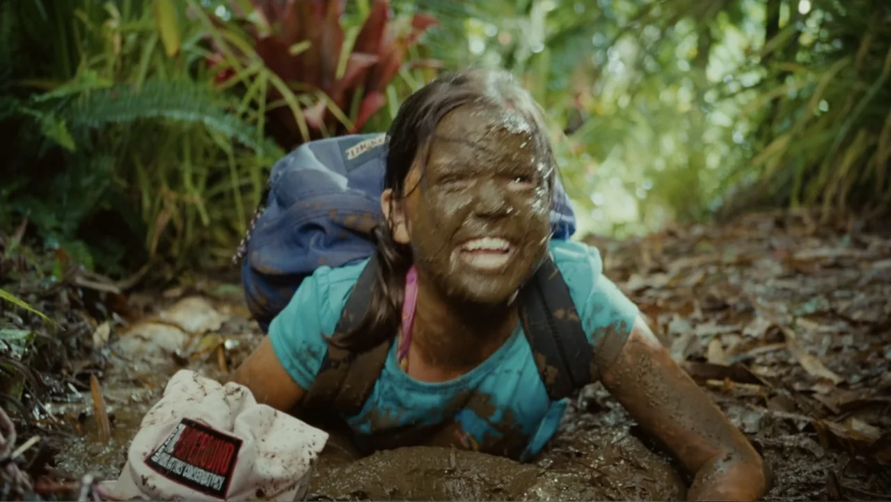 Just Go with It (2011) movie still: medium — A muddy path in a tropical jungle with dense foliage and large bromeliad plants. A teenage girl covered…