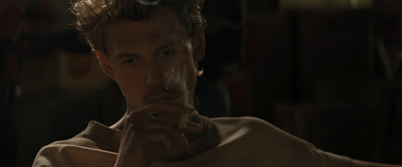 The Bikeriders (2023) movie still: closeup — A dimly lit room with muted earth tones and deep shadows. A man with disheveled hair holds a cigarette…