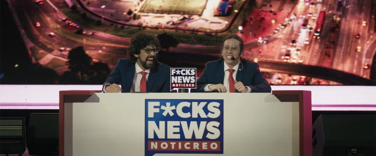 Making of: F*CKS NEWS (2025) movie still: medium — Two news anchors sit behind a white desk labeled with bold text in a studio. A large screen…