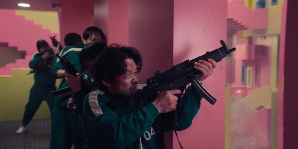 Squid Game (2024) movie still: medium — A group of people in teal tracksuits navigate a labyrinthine stairwell with pink walls. One man in the…