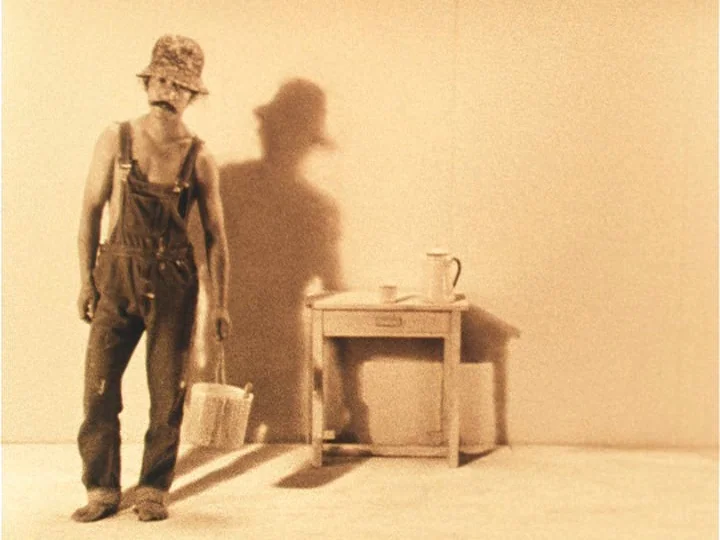 The Woman with Two Heads (1977) movie still: fullbody — A man stands in an empty, minimalist room with a sepia-toned finish. He holds a bucket while…