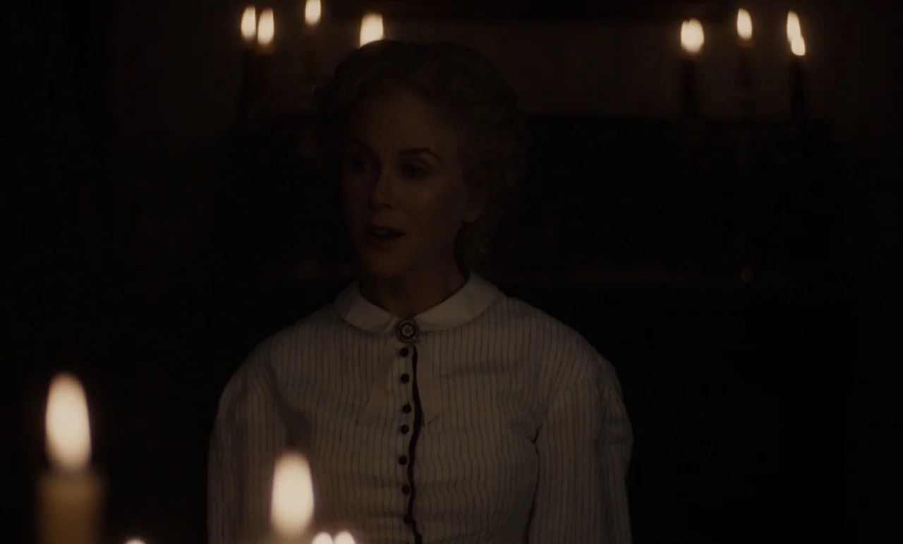 The Beguiled (2017) (2017) movie still: medium — A dimly lit room contains a woman seated at a wooden table at night. She wears a striped white…