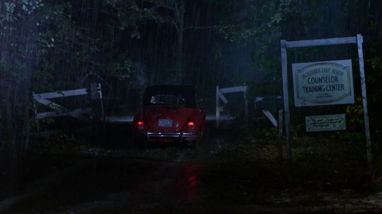Friday the 13th Part 2 (1981) movie still: wide — A red vintage car sits on a rainy dirt path at night near a gate. A weathered wooden sign…
