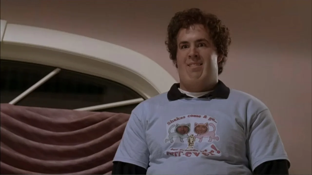 Just Friends (2005) movie still: medium — A man stands inside a room with plain walls and a window with curtains. He wears a graphic t-shirt and…