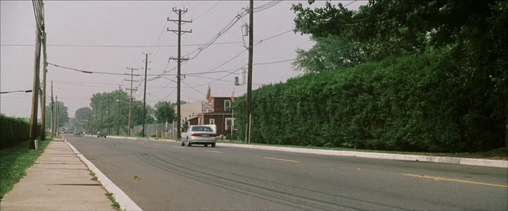Ocean’s Eleven (2001) movie still: wide — A suburban asphalt road runs past a dense hedge and overhead utility lines. A silver sedan travels away…