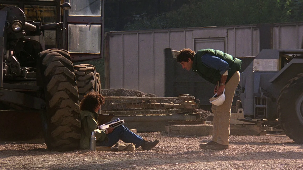 Flashdance (1983) movie still: fullbody — A gravel-covered construction site features heavy machinery and a metal shipping container. A man stands…