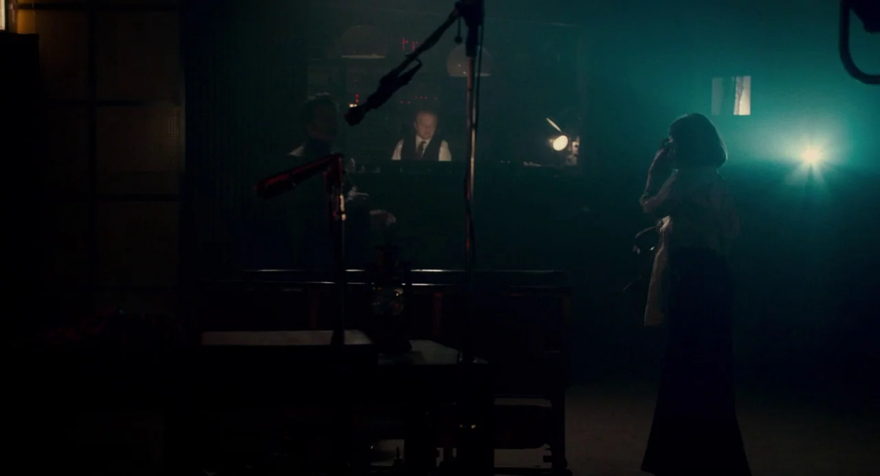 Berberian Sound Studio (2012) movie still: wide — A dimly lit room contains a broadcast booth with a man visible through glass. A woman stands in…
