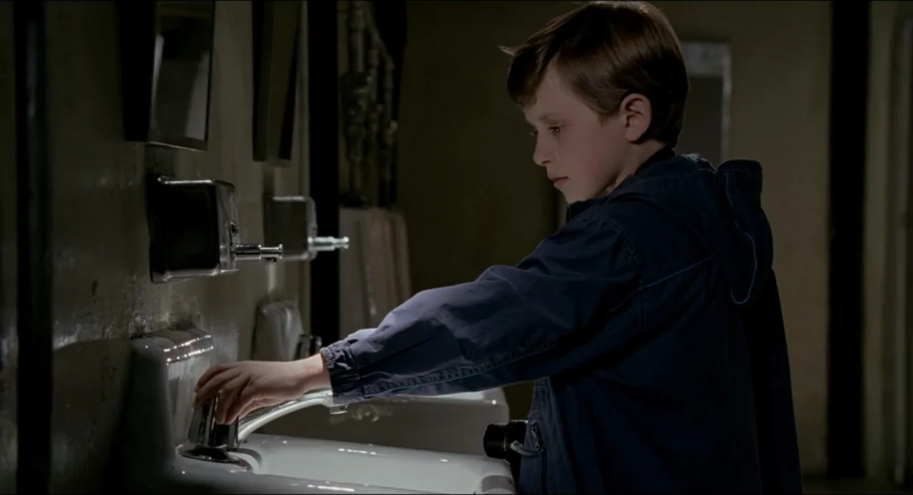 The Ring Two (2005) movie still: medium — A dimly lit public restroom features white ceramic basins and silver soap dispensers. A young boy in a…