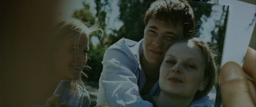 Next Door (2005) movie still: closeup — A handheld physical photograph shows two young women and a man together outdoors. The photograph displays a…