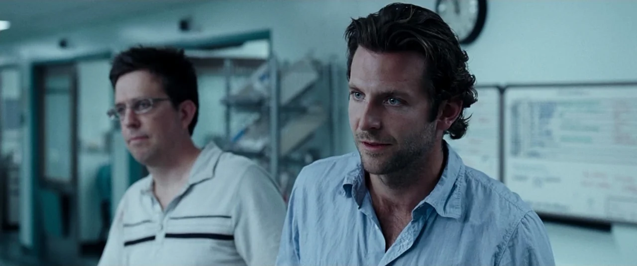 The Hangover (2009) movie still: medium — A hospital hallway features cool-toned walls, a wall clock, and white medical charts. Two men in casual…