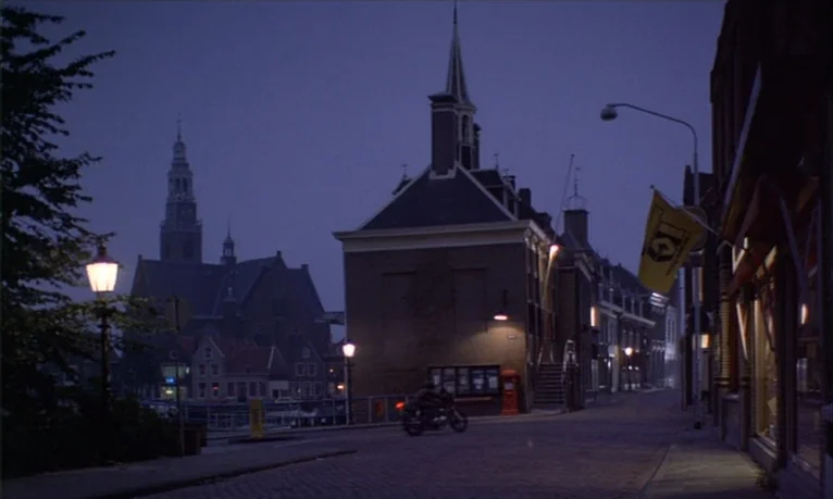 Spetters (1980) movie still: wide — A European town square at night features brick buildings and a tall church spire. A lone motorcycle travels down…