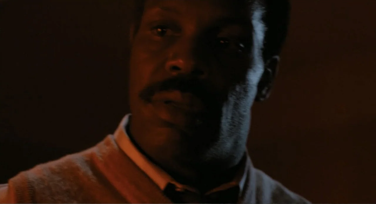 Lethal Weapon (1987) movie still: closeup — A dark indoor setting with limited illumination on a man's face. An older man gazes toward the side of…