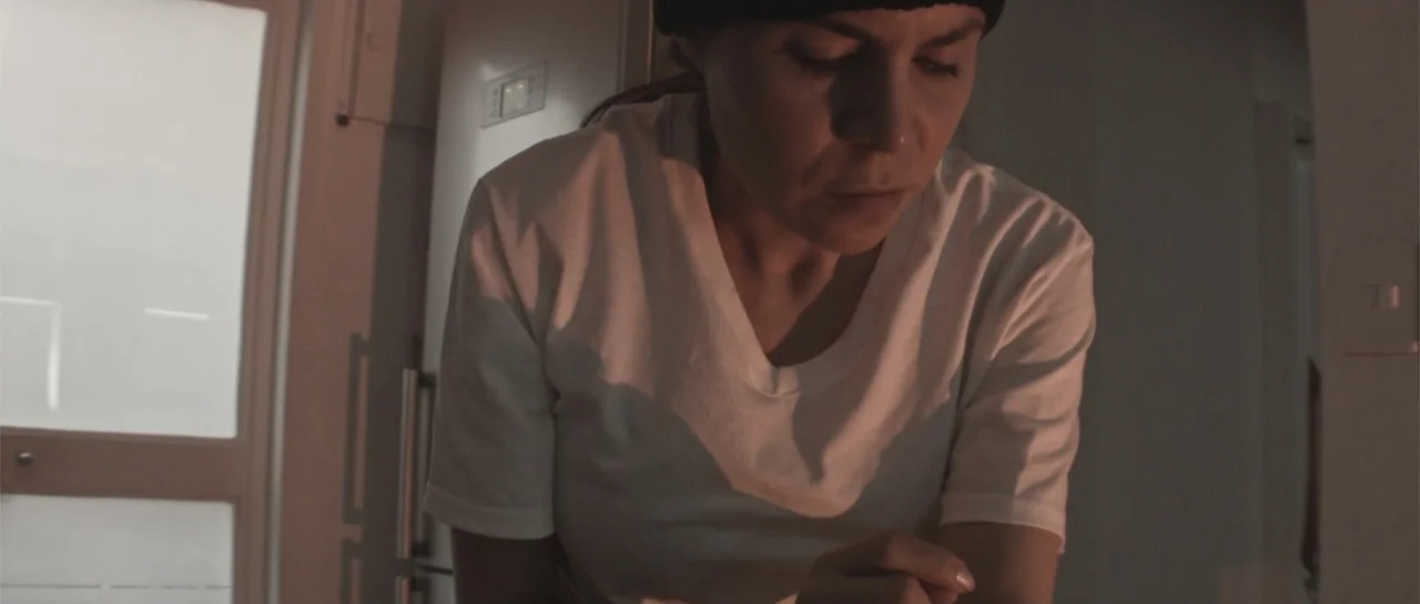 Amor Mutante (2019) movie still: medium — A dimly lit indoor space with a plain white door and white walls visible. A woman in a white t-shirt…