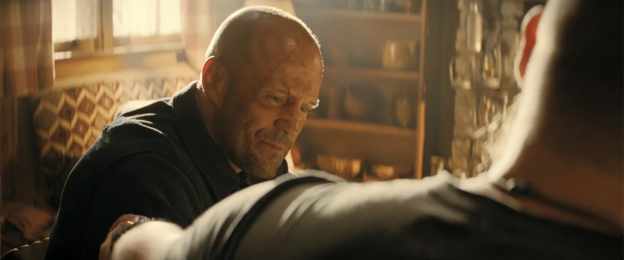A Working Man (2025) movie still: medium — A rustic cabin interior features wood paneled walls and shelves filled with kitchenware. A man with a…